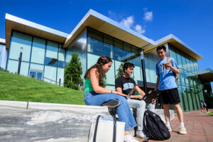 Three students sit in front of the Bloomberg Student Center, a large glass building
