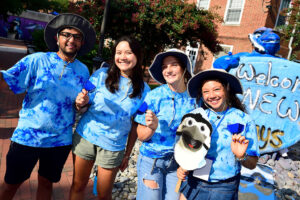 A group of students, smiling and wearing blue, standing in front of a sign that reads "Welcome, new Jays!"