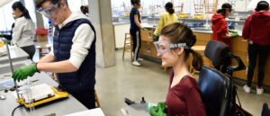 Two students wearing goggles and working in a lab; one of the students is seated in a wheelchair.