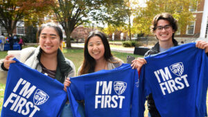 Three students hold up blue t-shirts that read "I'm First"