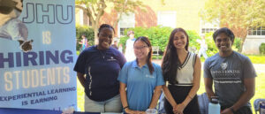 Four students standing at a table, next to a banner that reads "JHU is Hiring Students"